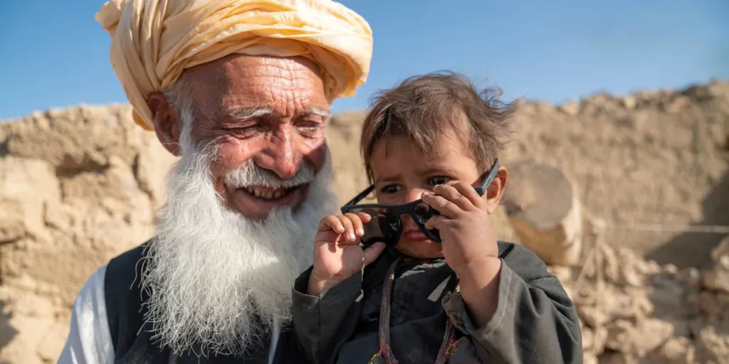 A boy puts on sunglasses while his grandfather holds him, smiling. In Afghanistan, UNHCR is providing earthquake-resilient houses for displaced families.